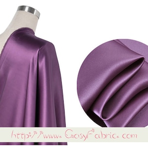 May include: A close-up of a purple satin fabric. The fabric is smooth and silky, with a slight sheen. It is draped over a mannequin and folded in a way that shows its texture.