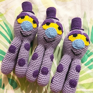 Puede incluir: Tres peluches de pulpo de color morado hechos a crochet con ojos amarillos, narices azules y sombreros morados. Los peluches tienen un patrón de lunares.