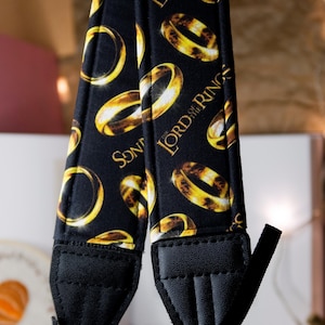 Può includere: Cinturino per fotocamera nero con un motivo ripetuto di anelli dorati e la scritta "LORD OF THE RINGS" in oro. Il cinturino ha estremità in pelle nera e un cinturino regolabile nero.