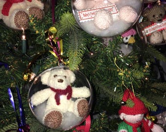 May include: A Christmas tree decorated with clear glass ornaments containing knitted toys. The ornaments feature a white teddy bear with a red scarf, a pink doll with a pink hat, a white teddy bear with a red scarf, a green and red elf, and a snowman.