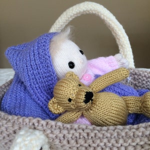 May include: A knitted basket with a purple hooded doll and a brown teddy bear inside. The basket has a white handle.