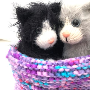 May include: Two knitted plush cat toys, one black and white and one grey, are nestled together inside a colourful knitted basket.