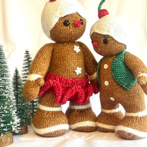 Könnte beinhalten: Zwei gestrickte Lebkuchenmännchen-Puppen. Eine Puppe trägt einen roten Rock, die andere eine grüne Weste. Beide haben weiße Hüte mit roten Kirschakzenten, schwarze Augen und weiße Schneeflocken-Details. Weihnachtsdekoration.