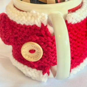 Puede incluir: Una taza de cer&aacute;mica blanca con una funda de punto roja y blanca. La funda presenta un cuerpo rojo con ribete blanco y un bot&oacute;n de madera que dice "Handmade with love". La taza contiene varios paquetes peque&ntilde;os.