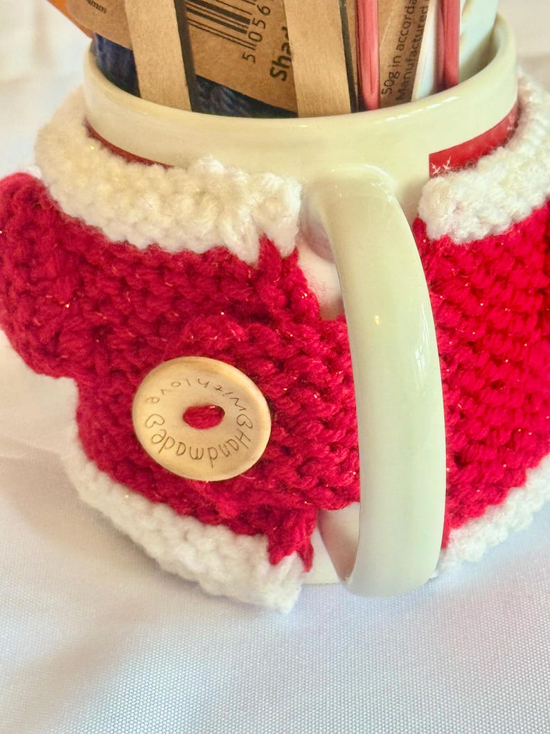 Puede incluir: Una taza de cer&aacute;mica blanca con una funda de punto roja y blanca. La funda presenta un cuerpo rojo con ribete blanco y un bot&oacute;n de madera que dice "Handmade with love". La taza contiene varios paquetes peque&ntilde;os.