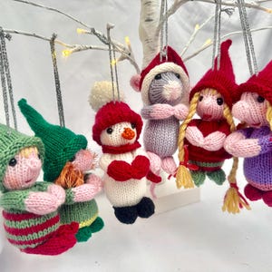 May include: A collection of six hanging knitted ornaments featuring two elves in green hats, a snowman with a red hat and scarf, a grey mouse in a red hat, and two figures with blonde yarn braids wearing red hats and colorful knitted clothing.