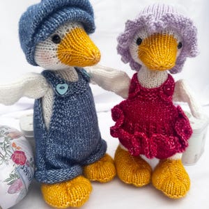 May include: Two knitted duck dolls. One wears blue dungarees and a matching cap. The other wears a red dress and a purple bonnet. Both ducks have yellow beaks and feet, and white wings.