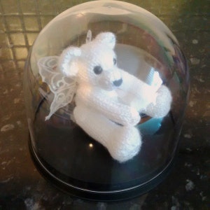 May include: A white knitted teddy bear with wings is displayed inside a clear glass dome. The bear is sitting with its wings spread out and its head tilted to the side. The dome is on a black base.