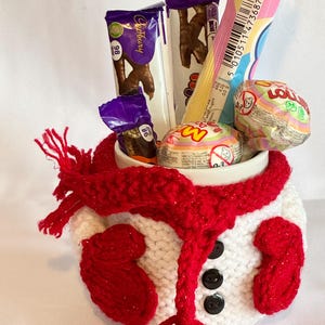 Puede incluir: Una taza de cer&aacute;mica blanca decorada como un mu&ntilde;eco de nieve, con una bufanda y mitones rojos. La taza est&aacute; llena de varios caramelos y barras de chocolate, incluyendo Cadbury. Botones negros est&aacute;n cosidos en la parte delantera.