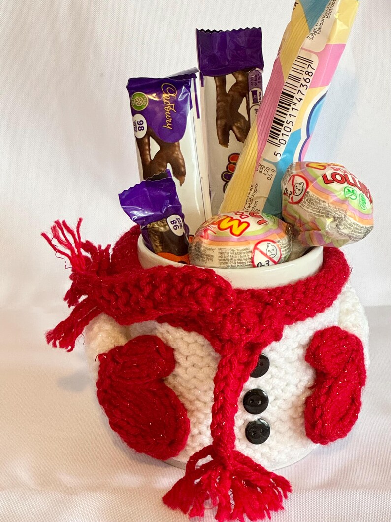 Puede incluir: Una taza de cer&aacute;mica blanca decorada como un mu&ntilde;eco de nieve, con una bufanda y mitones rojos. La taza est&aacute; llena de varios caramelos y barras de chocolate, incluyendo Cadbury. Botones negros est&aacute;n cosidos en la parte delantera.