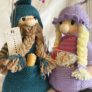 May include: Two knitted pincushions, one blue and one purple, with yarn, needles, and buttons. The blue pincushion has a beard and a hat, and the purple pincushion has pigtails and a hat. The pincushions are sitting on a white surface.