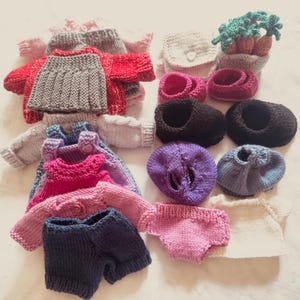 Puede incluir: Una colección de ropa de muñeca tejida a mano en varios colores. El surtido incluye suéteres, pantalones cortos, zapatos y sombreros. Los colores incluyen rojo, rosa, gris, azul y negro. Los artículos están dispuestos sobre una superficie blanca.