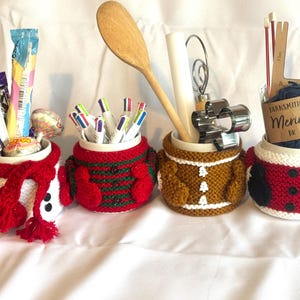 Puede incluir: Cuatro tazas de cer&aacute;mica con fundas de punto con dise&ntilde;os festivos. Las tazas contienen varios art&iacute;culos: caramelos, bol&iacute;grafos, utensilios de cocina e hilo. Las fundas incluyen un mu&ntilde;eco de nieve, un su&eacute;ter navide&ntilde;o, un hombre de jengibre y un dise&ntilde;o de Pap&aacute; Noel.