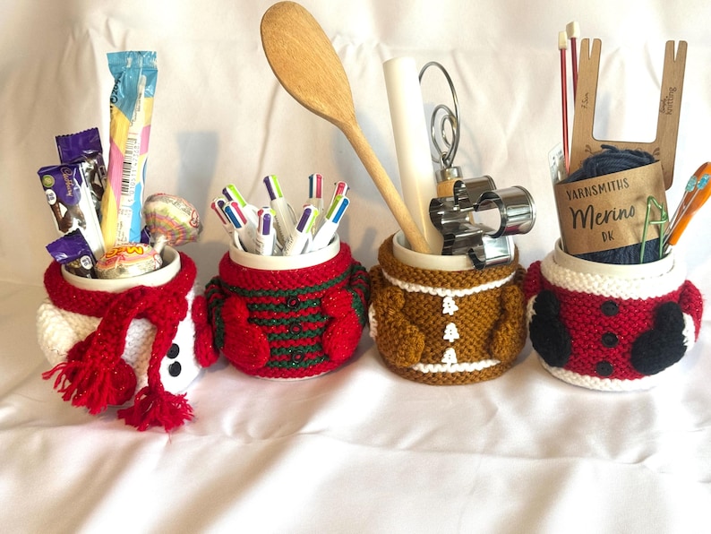 Puede incluir: Cuatro tazas de cer&aacute;mica con fundas de punto con dise&ntilde;os festivos. Las tazas contienen varios art&iacute;culos: caramelos, bol&iacute;grafos, utensilios de cocina e hilo. Las fundas incluyen un mu&ntilde;eco de nieve, un su&eacute;ter navide&ntilde;o, un hombre de jengibre y un dise&ntilde;o de Pap&aacute; Noel.