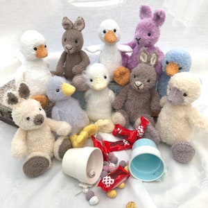 May include: A group of knitted animal toys, including bunnies, ducks, and a lamb, are arranged around two small ceramic pots and a pile of colorful candy eggs. The toys are all white, gray, brown, purple, and blue.