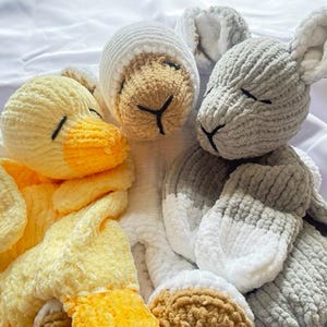 Puede incluir: Tres animales de peluche tejidos a crochet: un pato amarillo, una oveja marrón y blanca y un conejito gris y blanco. Los animales están todos acostados y tienen los ojos cerrados.