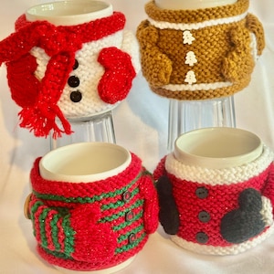 Puede incluir: Cuatro tazas de cer&aacute;mica con fundas de punto con dise&ntilde;os festivos. Una taza est&aacute; vestida como un mu&ntilde;eco de nieve con bufanda y mitones rojos. Otra se asemeja a un hombre de jengibre. Las otras dos tazas est&aacute;n vestidas como Pap&aacute; Noel.