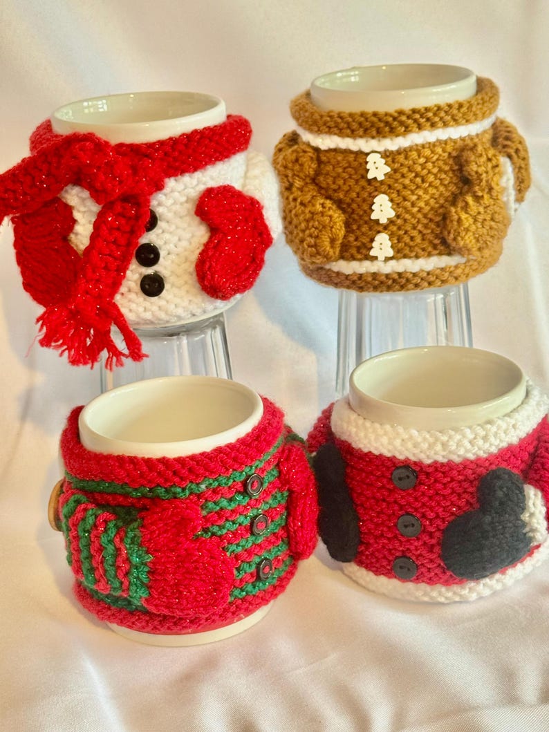 Puede incluir: Cuatro tazas de cer&aacute;mica con fundas de punto con dise&ntilde;os festivos. Una taza est&aacute; vestida como un mu&ntilde;eco de nieve con bufanda y mitones rojos. Otra se asemeja a un hombre de jengibre. Las otras dos tazas est&aacute;n vestidas como Pap&aacute; Noel.