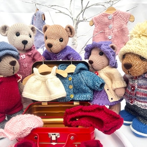 May include: A collection of five knitted teddy bears wearing various outfits including hats, sweaters, and overalls. A red metal suitcase sits in the centre, containing a white dress, a blue cardigan, and a red bonnet, with extra clothing hanging above.