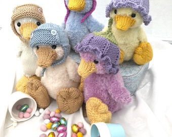 May include: Five knitted duck plushies wearing colorful hats. The ducks are sitting on a vintage suitcase with a blue and white pattern. The ducks are surrounded by chocolate Easter eggs.