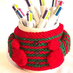 Puede incluir: Una taza de cer&aacute;mica blanca llena de bol&iacute;grafos coloridos, envuelta en una funda de punto roja y verde. La funda presenta un patr&oacute;n de rayas, tres botones negros y peque&ntilde;os detalles en forma de mitones a cada lado. Los bol&iacute;grafos tienen puntas multicolores.