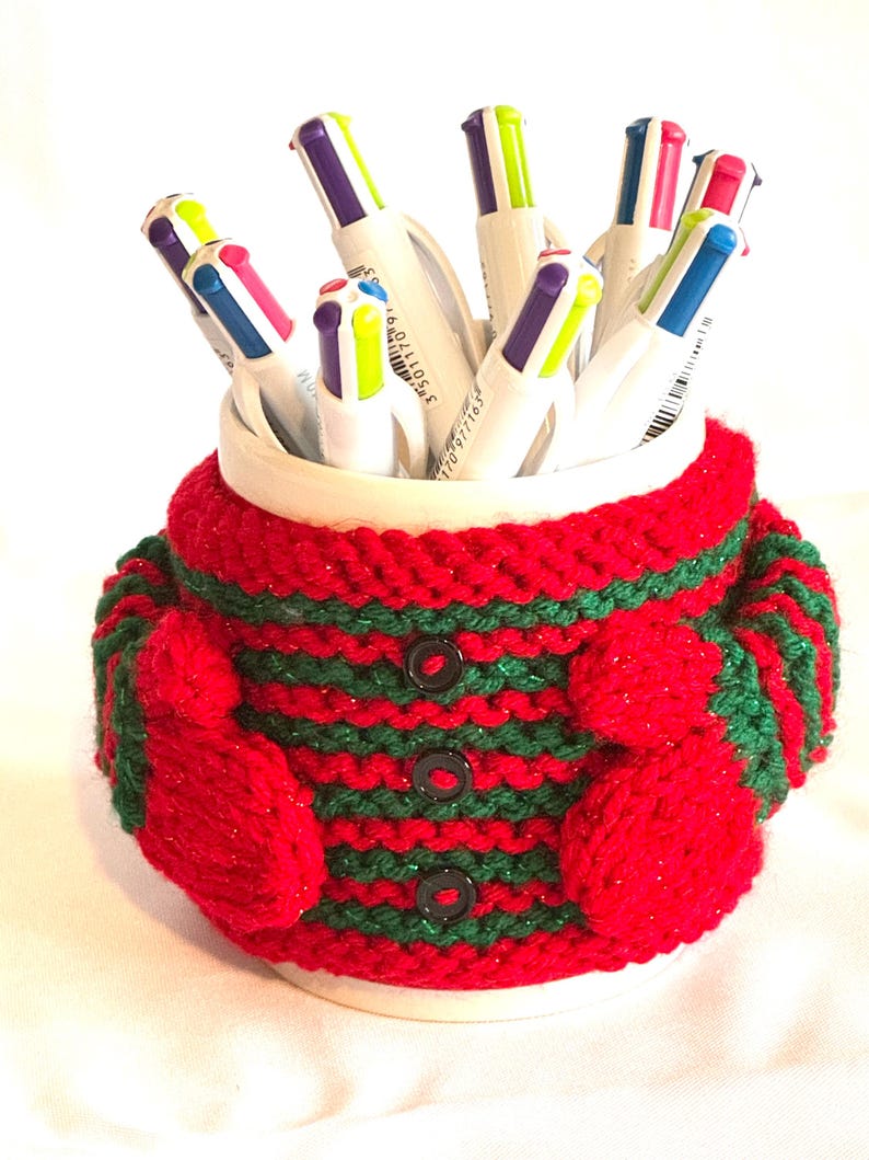 Puede incluir: Una taza de cer&aacute;mica blanca llena de bol&iacute;grafos coloridos, envuelta en una funda de punto roja y verde. La funda presenta un patr&oacute;n de rayas, tres botones negros y peque&ntilde;os detalles en forma de mitones a cada lado. Los bol&iacute;grafos tienen puntas multicolores.