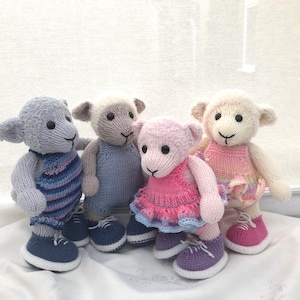 Puede incluir: Cuatro corderos de peluche tejidos que llevan atuendos de diferentes colores. Los corderos están de pie sobre una superficie blanca. El cordero de la izquierda lleva un atuendo a rayas azul y rosa, el cordero del medio lleva un atuendo azul, el cordero de la derecha lleva un atuendo rosa y el cordero de atrás lleva un atuendo rosa y amarillo.