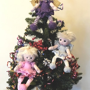 May include: Three knitted angel dolls with blonde hair and wings are decorating a Christmas tree. The angels are wearing purple, pink, and gray dresses with white stripes on their legs. The tree is decorated with red berries, gold and silver ribbon, and a silver star.