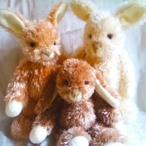 May include: Three plush bunny rabbits, one brown, one light brown, and one white. The bunnies are sitting together on a white surface.