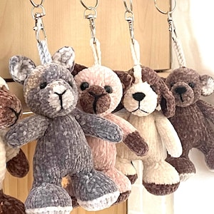 May include: Five crocheted plush animal keychains in various colors and styles. The keychains include a brown and white sheep, a gray and white dog, a pink and brown dog, a white and brown dog, and a brown dog.
