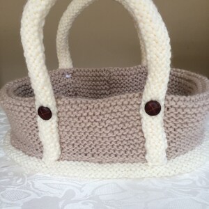 May include: A knitted beige and white basket with two handles and brown buttons. The basket is sitting on a white tablecloth.