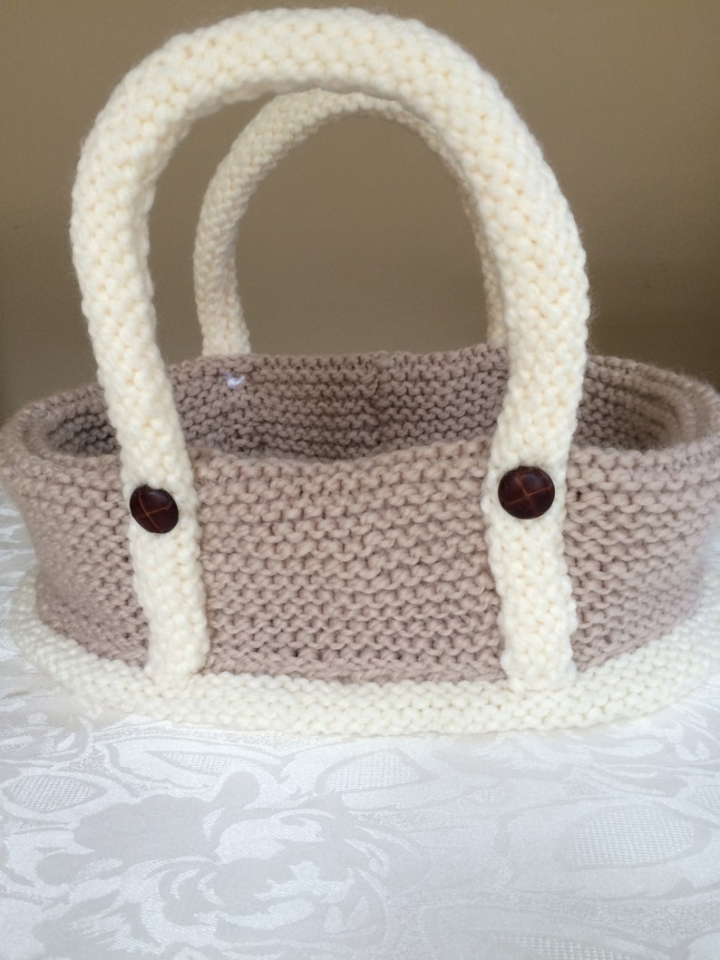 May include: A knitted beige and white basket with two handles and brown buttons. The basket is sitting on a white tablecloth.