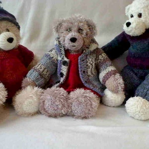 May include: Three plush teddy bears wearing knitted clothing. The bear on the left is wearing a red and white knitted sweater and a blue and white knitted hat. The bear in the middle is wearing a brown, white, and purple knitted sweater. The bear on the right is wearing a purple and blue knitted sweater.