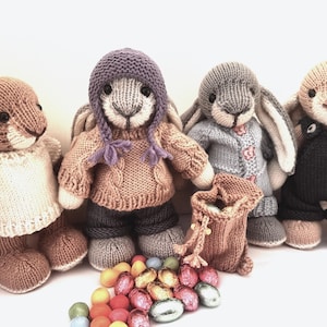 May include: Four knitted bunny rabbits wearing different outfits, including a purple hat, a blue sweater, a brown sweater, and overalls. The bunnies are surrounded by colourful chocolate Easter eggs.