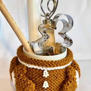 Puede incluir: Un recipiente de cer&aacute;mica con un dise&ntilde;o de hombre de jengibre. El recipiente est&aacute; envuelto en un acogedor tejido de ganchillo marr&oacute;n y blanco con tres botones en forma de &aacute;rbol. En el interior hay una cuchara de madera, un batidor de metal y un cortador de galletas de hombre de jengibre.