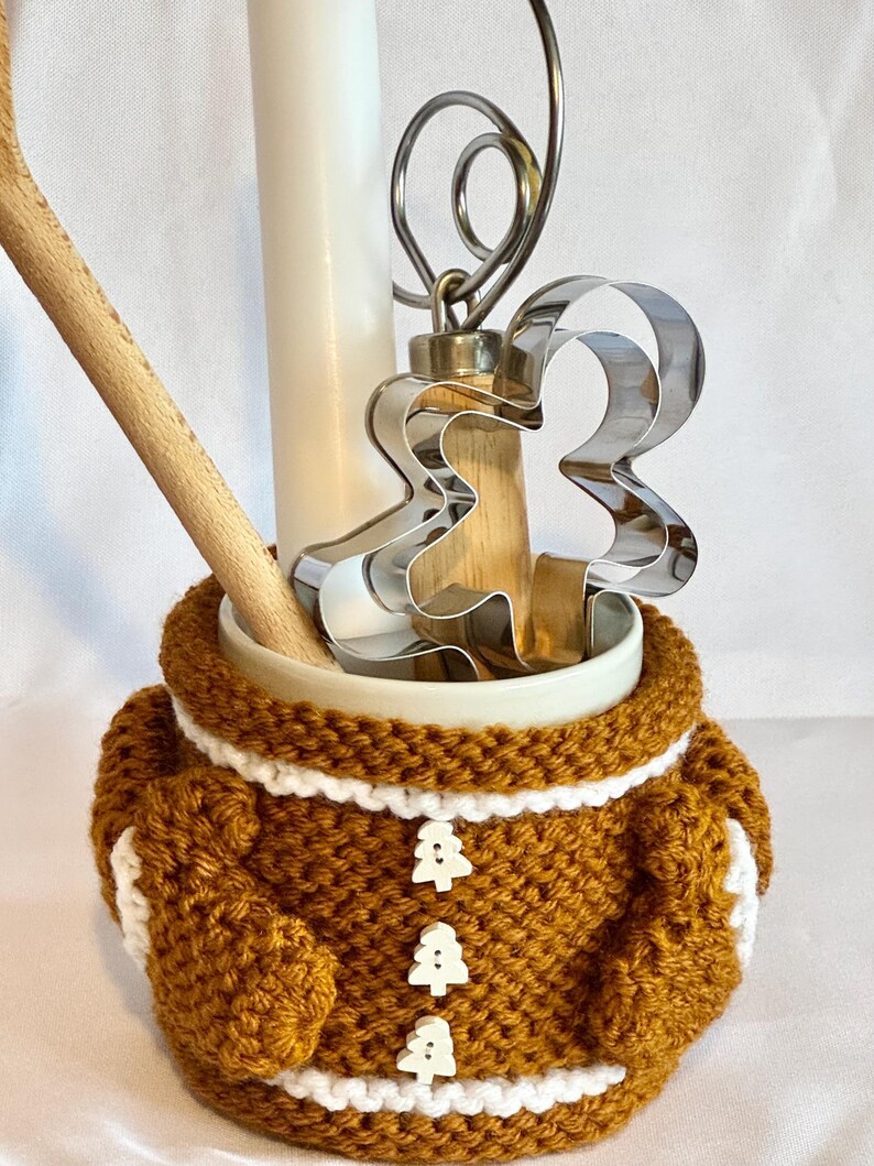 Puede incluir: Un recipiente de cer&aacute;mica con un dise&ntilde;o de hombre de jengibre. El recipiente est&aacute; envuelto en un acogedor tejido de ganchillo marr&oacute;n y blanco con tres botones en forma de &aacute;rbol. En el interior hay una cuchara de madera, un batidor de metal y un cortador de galletas de hombre de jengibre.