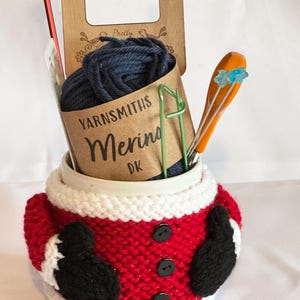 Puede incluir: Un kit de tejido navide&ntilde;o en un acogedor tema de Santa. El kit incluye lana azul marino, agujas de tejer, un calibrador de madera y un sujetador de puntadas. El acogedor es rojo con adornos blancos, botones negros y mitones negros.