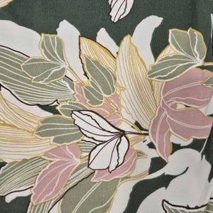 May include: Close-up of a fabric with a floral pattern. The design features large leaves and flower shapes in shades of white, green, pink, and yellow. The background is a dark green color.