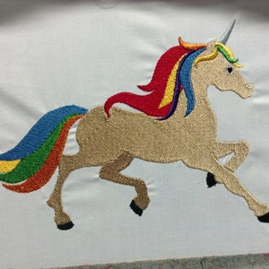 Puede incluir: Diseño de unicornio bordado con melena y cola de arcoíris. El unicornio es de color beige con un cuerno plateado y pezuñas negras. La melena y la cola presentan colores vibrantes como rojo, naranja, amarillo, verde y azul.