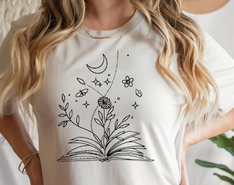 Puede incluir: Una camiseta blanca con un dibujo en línea negra de un libro abierto con flores, una luna creciente, estrellas y mariposas que crecen de él.