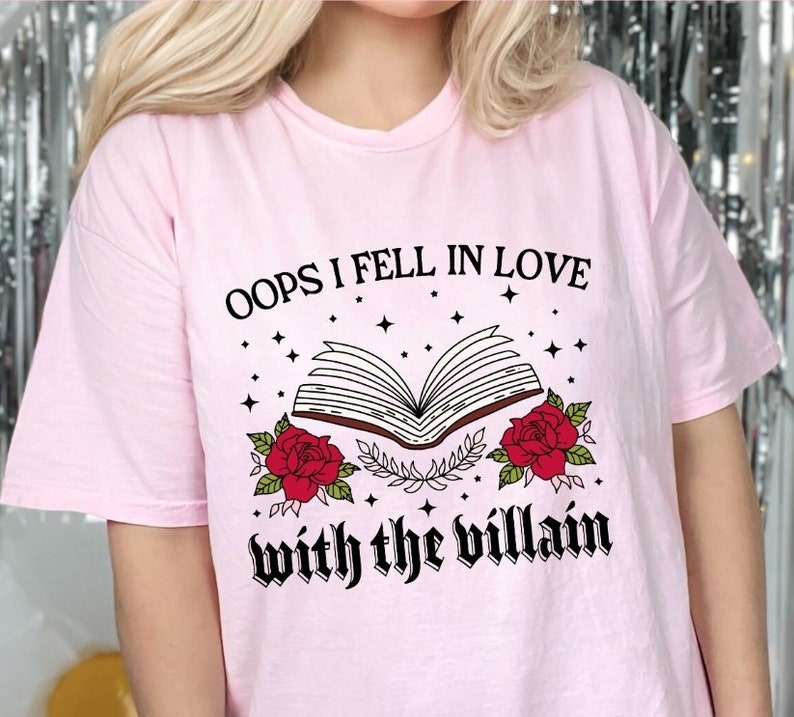 Może przedstawiać: R&oacute;żowa koszulka z czarnym nadrukiem otwartego książki z czerwonymi r&oacute;żami i tekstem "Oops I fell in love with the villain".