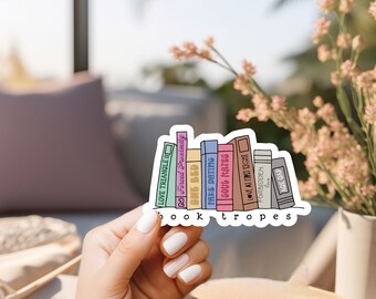 Puede incluir: Una pegatina con una ilustración colorida de una estantería con libros titulados "Love Triangle II", "Forced Proximity", "One Bed", "Fake Dating", "Soul Mates", "Love at First Sight", "Forbidden Love" y "Age Gap". La pegatina dice "book tropes" en la parte inferior.
