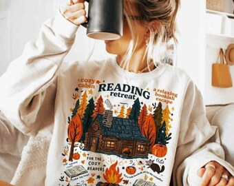 May include: A cream-coloured sweatshirt featuring a colourful graphic of a cabin in a forest, with the words "Reading Retreat" and other text. The design includes a campfire, books, and pumpkins. A person is holding a black mug.