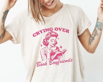 May include: A white t-shirt with a pink graphic of a woman reading a book and crying. The text on the shirt reads "Crying over Book Boyfriends".