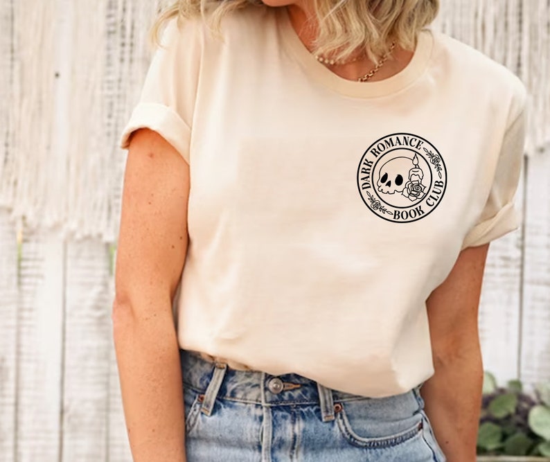 Peut inclure: Un t-shirt cr&egrave;me avec un motif noir repr&eacute;sentant un cr&acirc;ne avec une rose et une bougie &agrave; l'int&eacute;rieur d'un cercle avec le texte "Dark Romance Book Club".