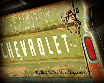 May include: A vintage green Chevrolet pickup truck with rust and faded paint. The truck has a chain and hook attached to the tailgate. The word "Chevrolet" is written in white letters on the tailgate.