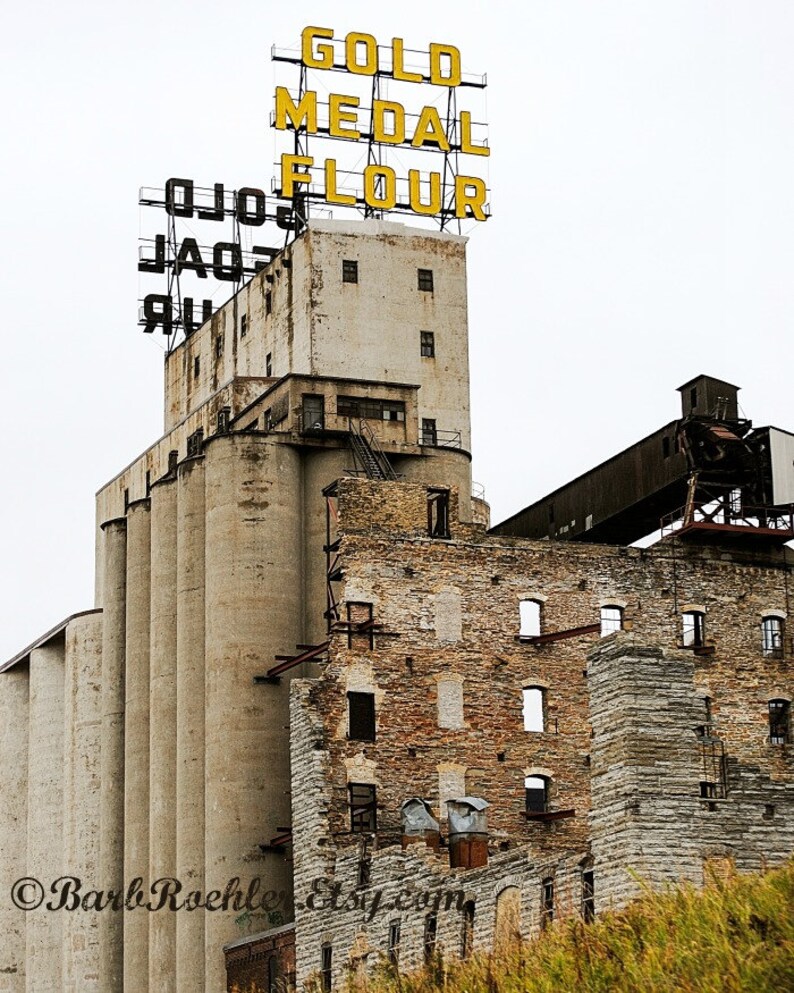 May include: A weathered brick building with a large sign that reads "GOLD MEDAL FLOUR" in yellow letters. The building is a grain silo with a series of large, cylindrical silos.