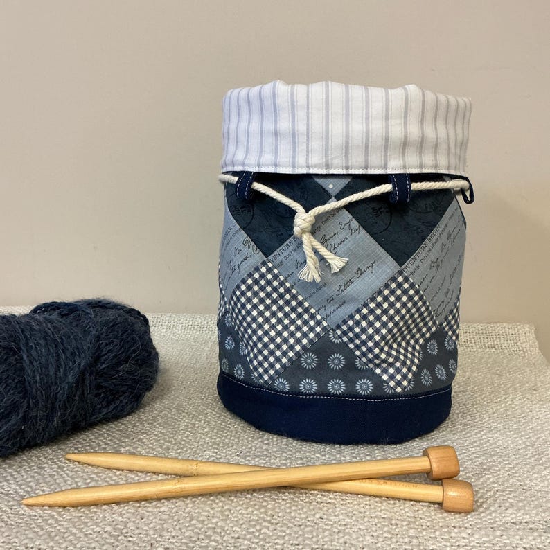 May include: A fabric yarn storage bag with a drawstring closure. The bag has a patchwork design in shades of blue and white, with a striped top. A ball of blue yarn and two wooden knitting needles are next to the bag.