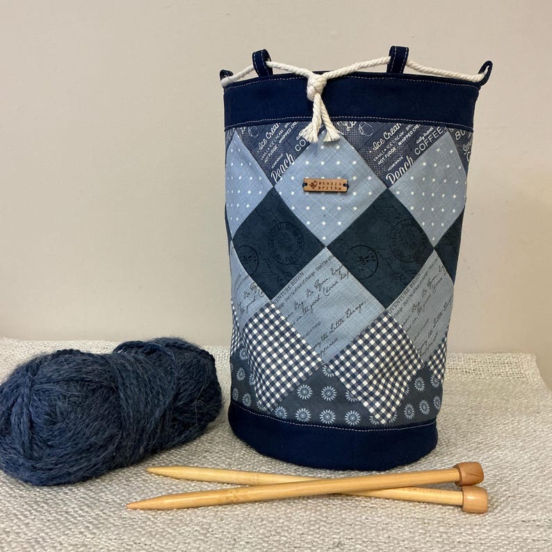 May include: A navy blue fabric yarn storage bag with a drawstring closure. The bag has a patchwork design with various blue patterns, including diamonds, polka dots, and floral prints. A ball of blue yarn and two wooden knitting needles are in the foreground.