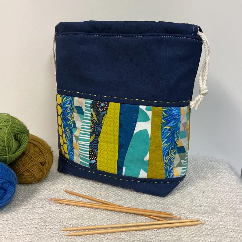 May include: A navy blue drawstring yarn bag with a colourful patchwork design. The patchwork includes vertical strips of blue, green, and yellow patterned fabric. Knitting needles and yarn balls are in the foreground.